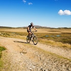 A biker is cycling along a rough track in the Australian countryside on a sunny day.