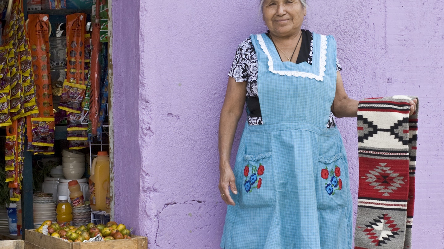 Smiling indigenous Mexican woman displaying a handwoven Zapotec rug outside a shop in Santa Ana del Valle.