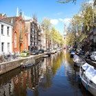 One of Amsterdam's central canals in the summer months. Boats full of people move through the water while the surrounding streets are thronged with people and bicycles.