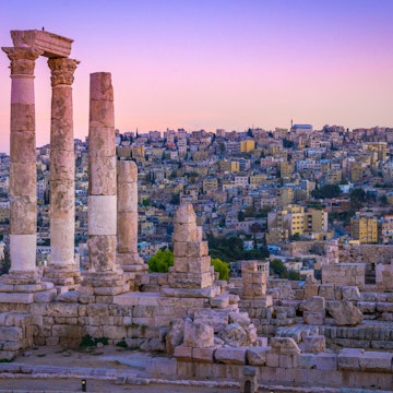 Several Roman columns stand tall and alone atop a hill, with the city in the distance as a backdrop; the sky is an incredible shad of pink and purple; Free things to do in Amman, Jordan