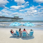 Family relaxing on  the beautiful beach, People enjoying summer vacation by the ocean. Family sitting under beach umbrella.  Cloudy sky and pier in the background. Folly Beach, South Carolina USA. ; Shutterstock ID 1428607988; your: Sloane Tucker; gl: 65050; netsuite: Online Editorial; full: Free Things South Carolina Article